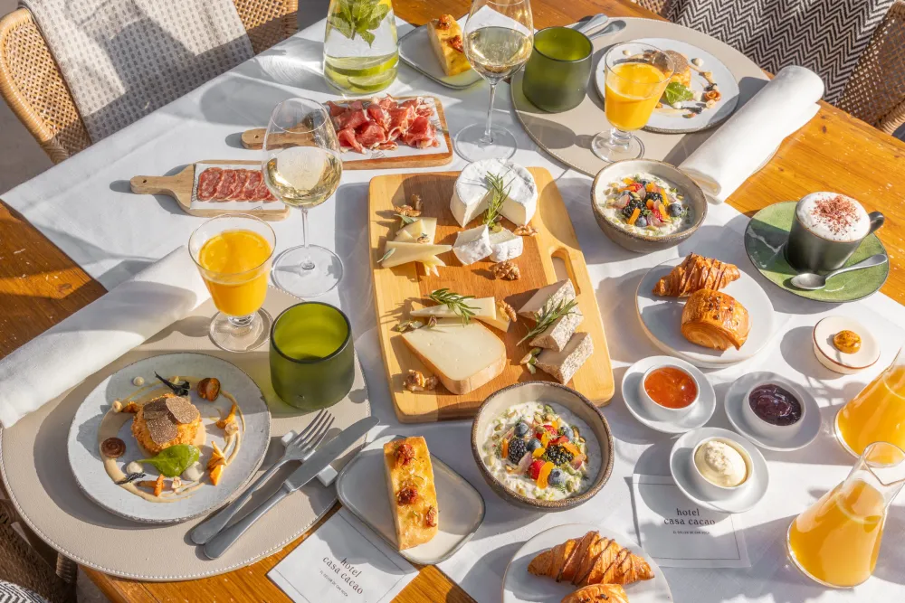 Mesa de desayuno de hotel con quesos, bollería, zumos y platos calientes al sol