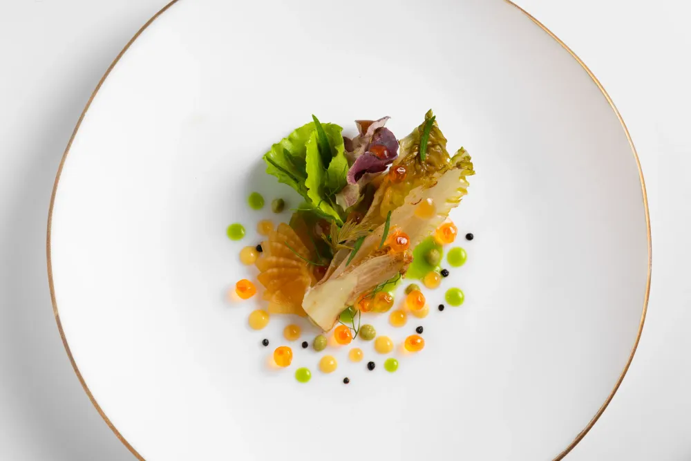 Plato blanco de borde dorado con una hoja de lechuga asada y perlas de salsa naranjas y verdes, visto desde arriba